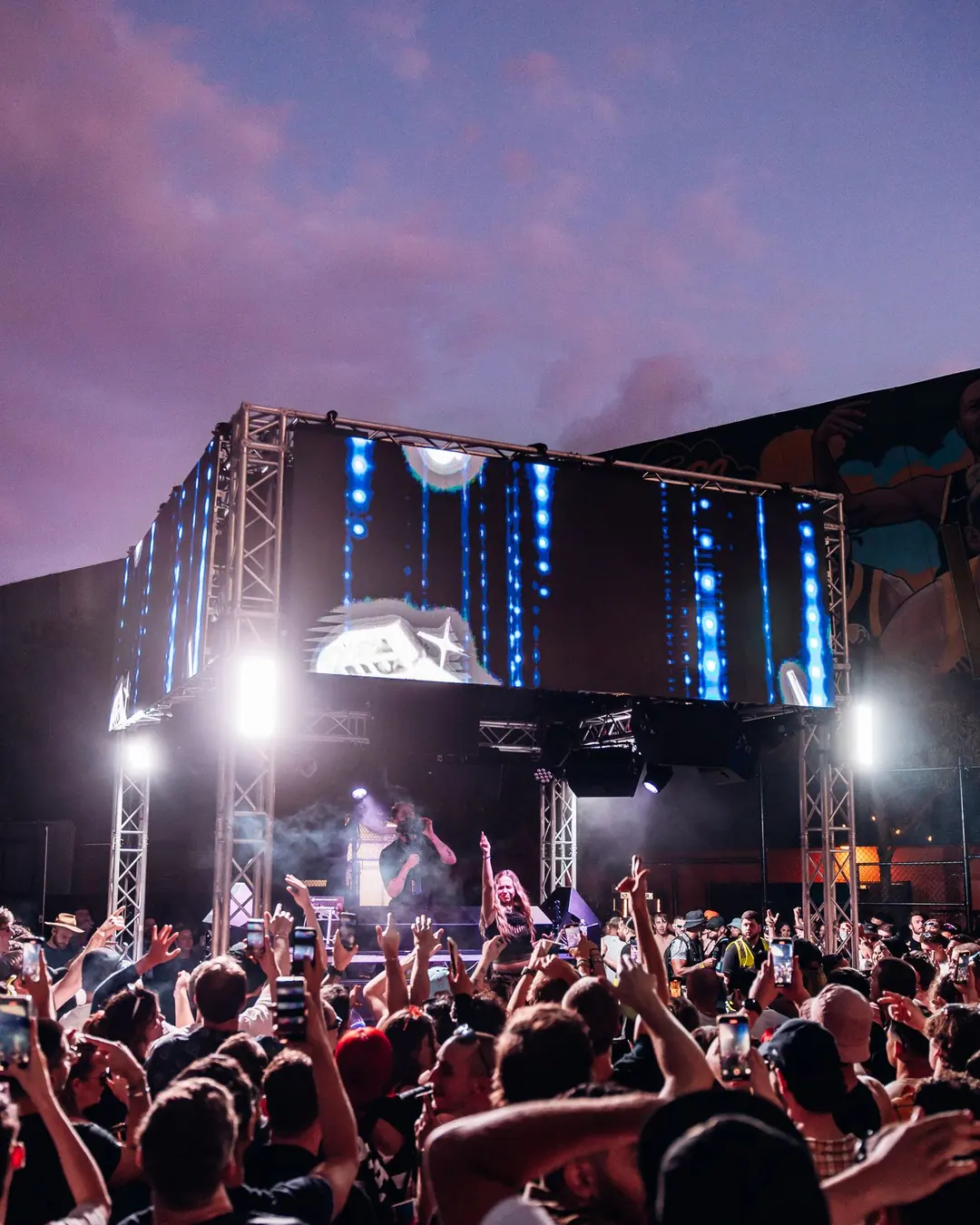 DnB Allstars: Sydney: Lit stage under a pink dusk sky with crowd hands up, outdoor peak