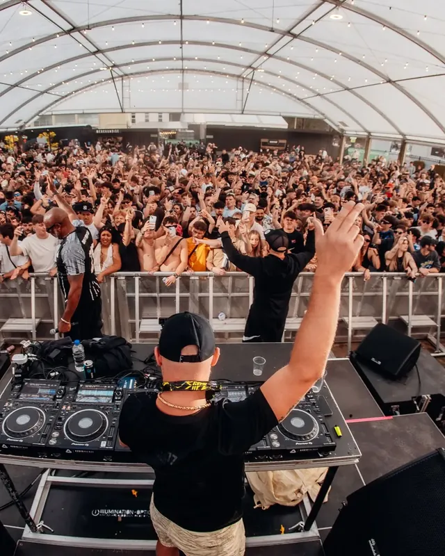 DnB Allstars: Wellington: DJ arm up facing huge tent crowd, wide fisheye