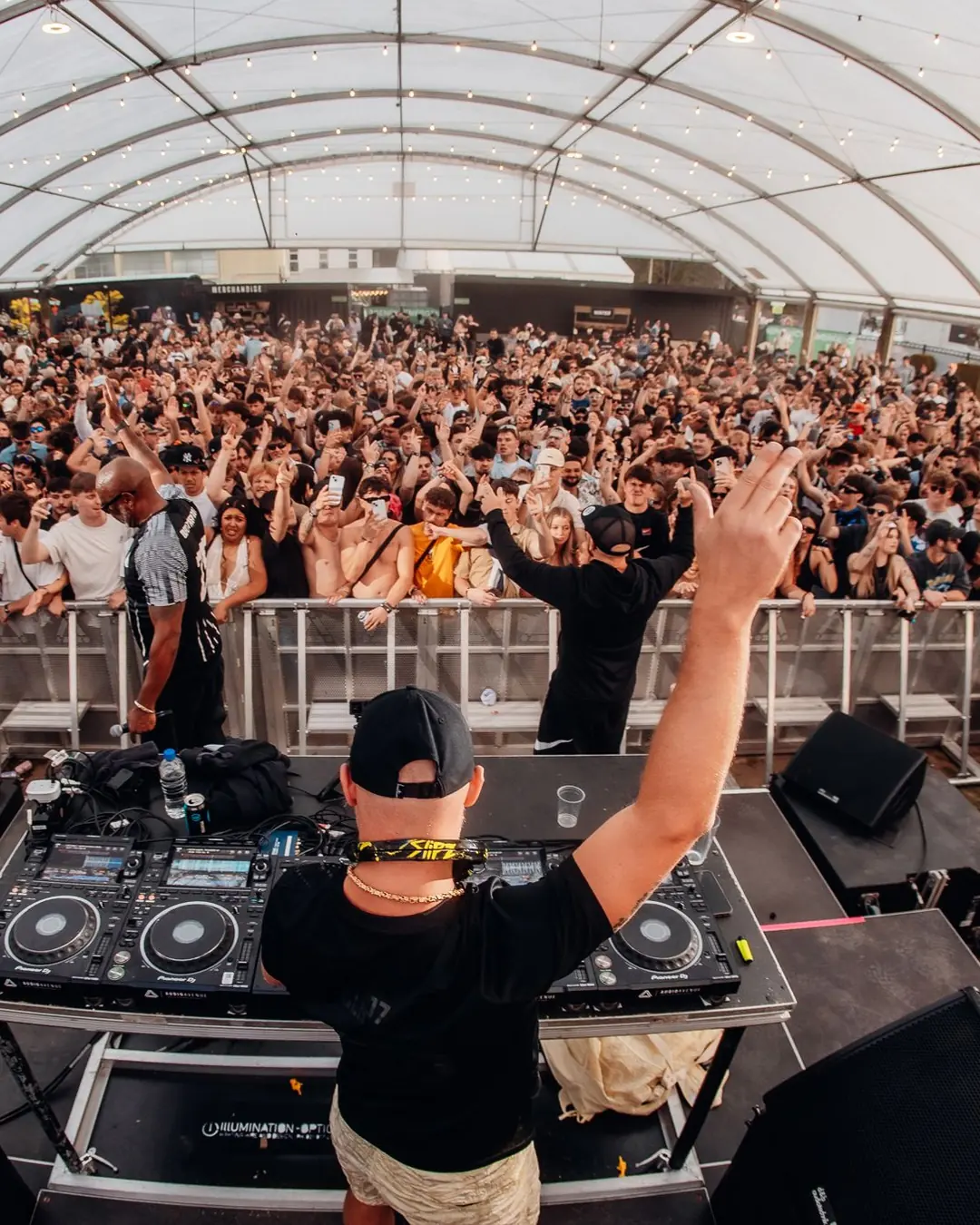 DnB Allstars: Wellington: DJ arm up facing huge tent crowd, wide fisheye