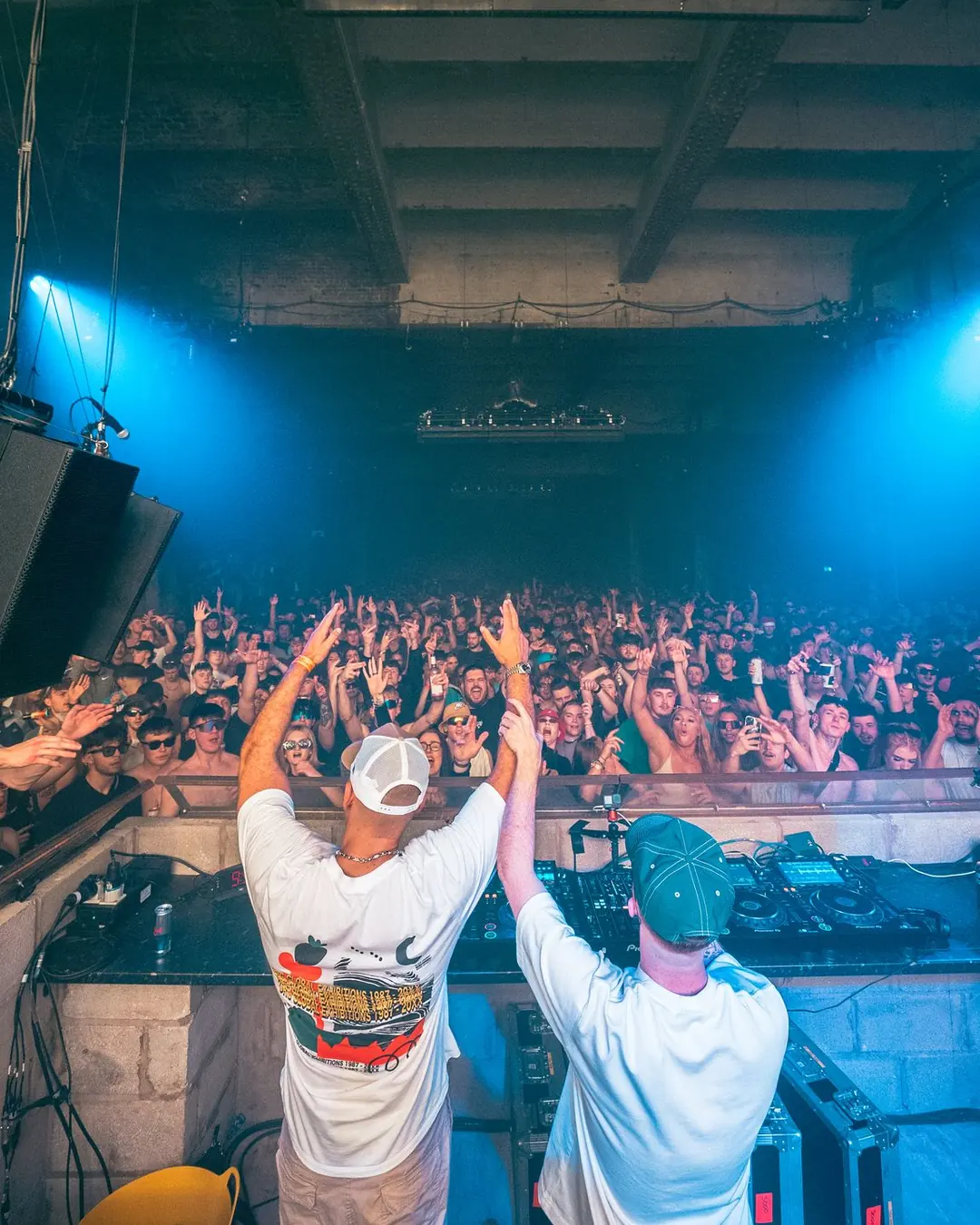 DnB Allstars x Warehouse Project: Manchester: Two artists arms up facing packed Depot crowd, blue lights